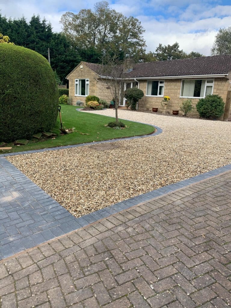 A single-story house with tan brick walls and a neatly landscaped front yard, featuring expert garden design, a paved driveway bordered by gravel, trimmed hedges, green grass, and small potted plants near the entrance.