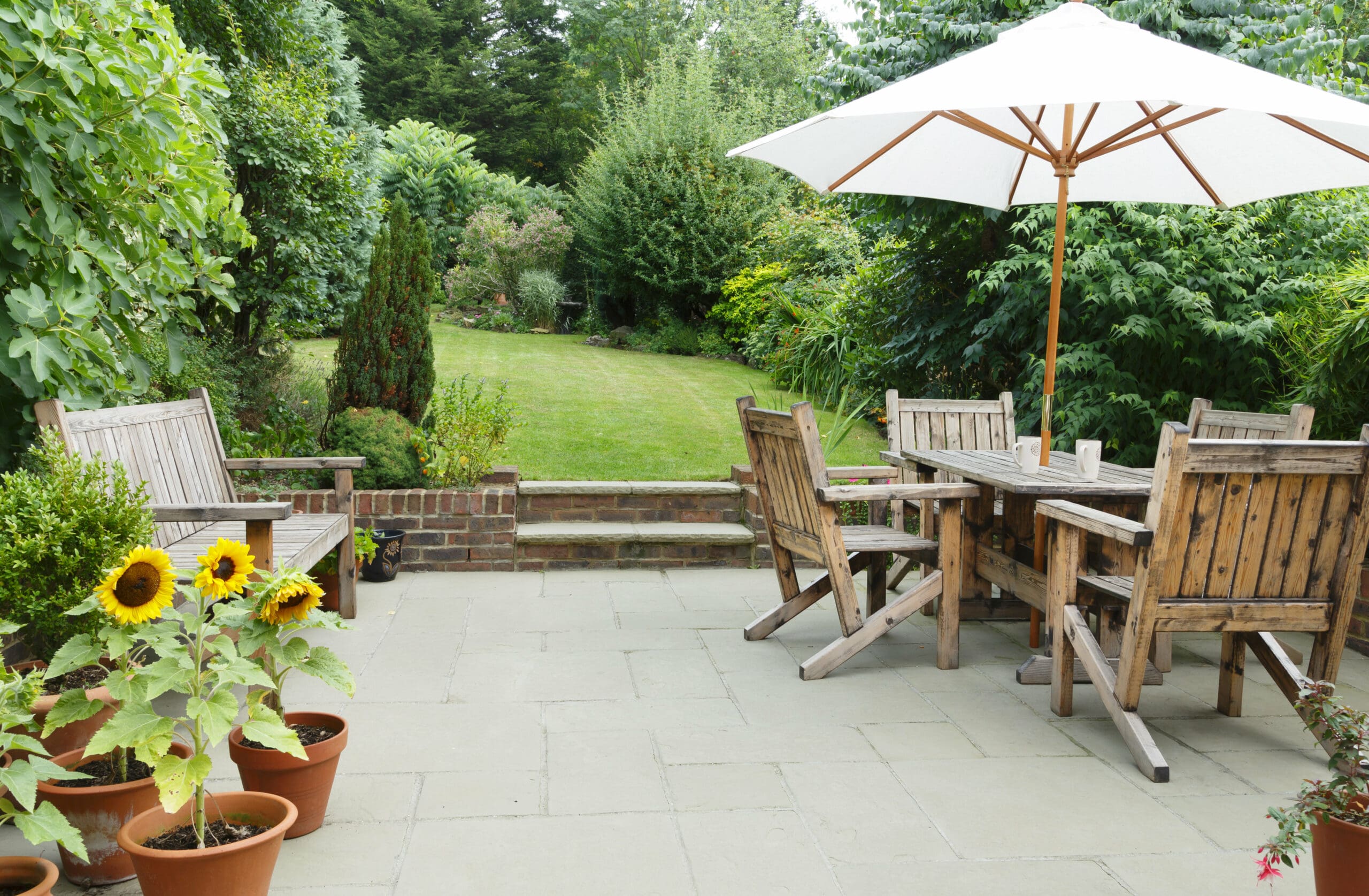 A patio with wooden benches and a dining table set under a large white umbrella, surrounded by potted plants and sunflowers, overlooks a lush green garden crafted by an expert landscaper, with trees and shrubs framing the serene space.