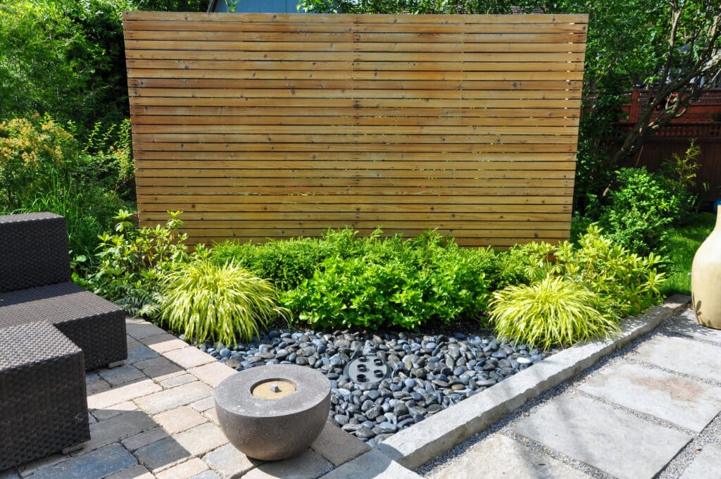 A modern backyard garden, crafted by an expert landscaper, features a wooden privacy screen, lush green shrubs, ornamental grasses, a small pebble bed, a round stone water fountain, and outdoor wicker furniture on a stone patio.